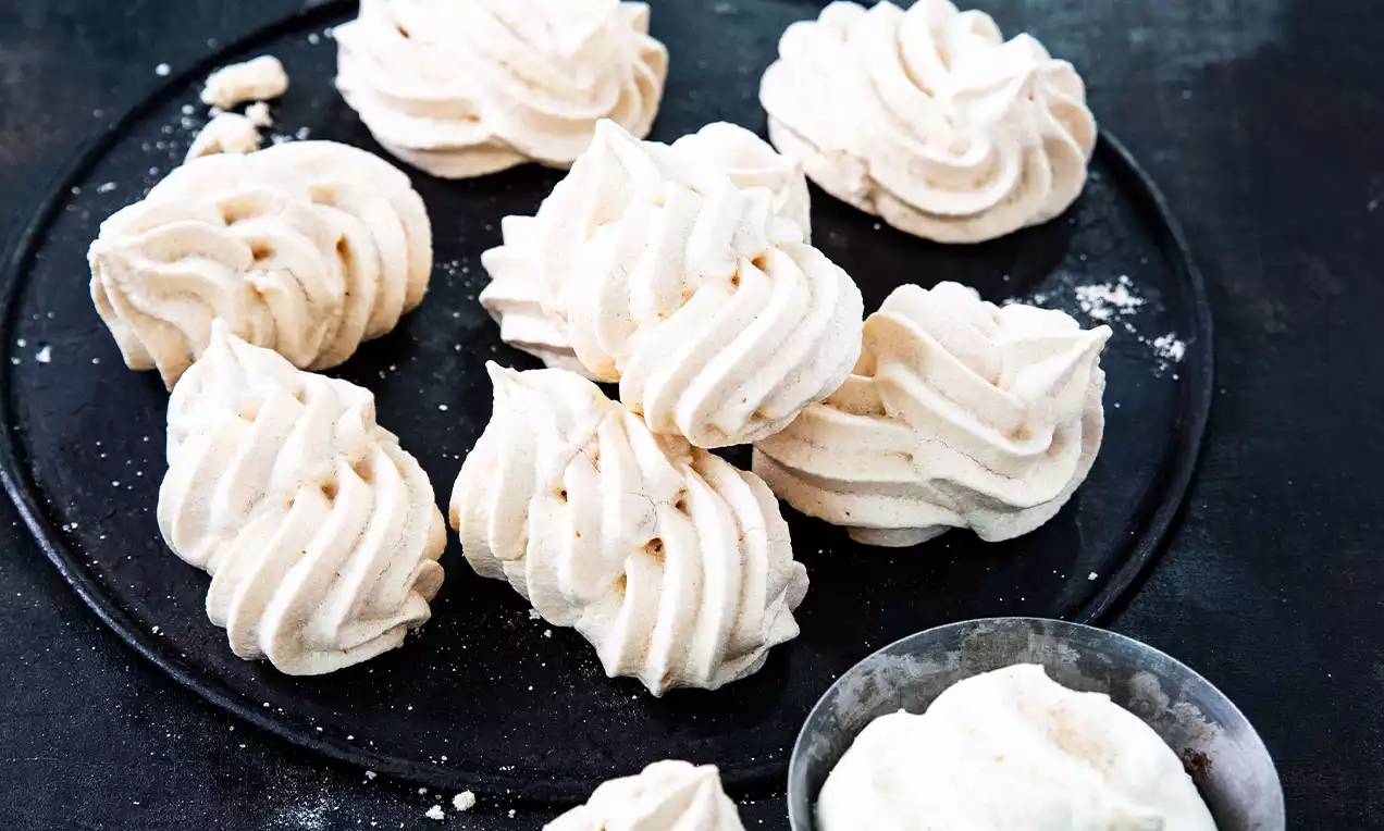 Meringhe bianche a forma di piccoli vortici arricciati, dalla superficie leggermente croccante, disposte su un piatto scuro con briciole sparse