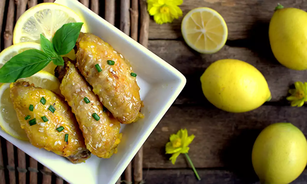 Golden roasted chicken wings with a glossy glaze, garnished with green chives and basil, served in a white dish with lemon slices