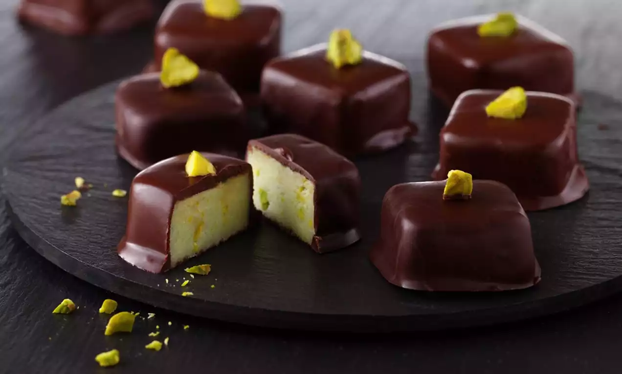 Square marzipan treats coated in glossy dark chocolate, topped with vibrant green pistachio pieces, arranged on a dark slate platter