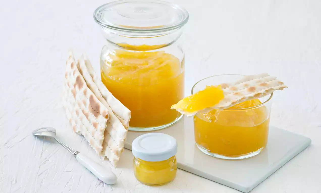 Sklenice sytě žlutého Piña Colada džemu s hladkou texturou, podávaný s křupavými suchary a ozdobený plátkem čerstvého ovoce na bílém pozadí