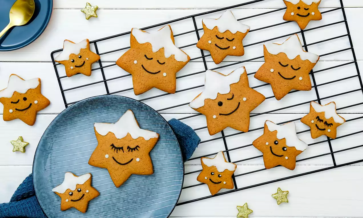 Goudbruine stervormige koekjes met witte glazuurdecoratie en geschilderde gezichten op een zwart rek, een blauw bord en een witte achtergrond