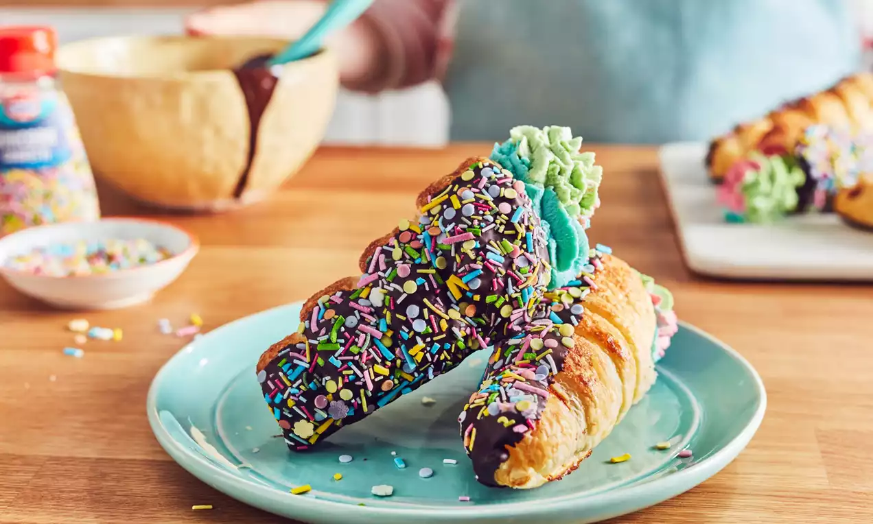 Golden cream horns with chocolate-dipped ends covered in colourful sprinkles, green and blue frosting, arranged on a light blue plate