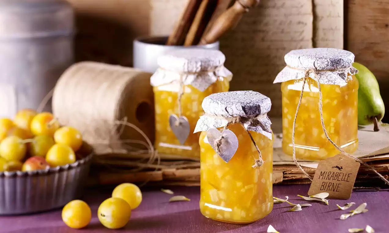 Tri sklenené poháre s jasnožltým mirabelkovo-hruškovým džemom, zakryté papierovými viečkami s motúzom a dekoratívnymi štítkami
