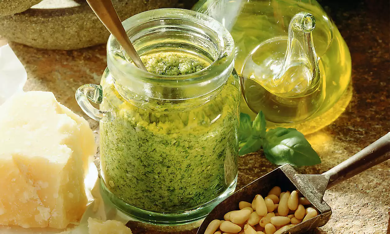 Grünes Pesto mit cremiger Textur in einem kleinen Glas mit Löffel, daneben Olivenöl in Glaskaraffe, Pinienkerne und Parmesanstück auf rustikalem Hintergrund