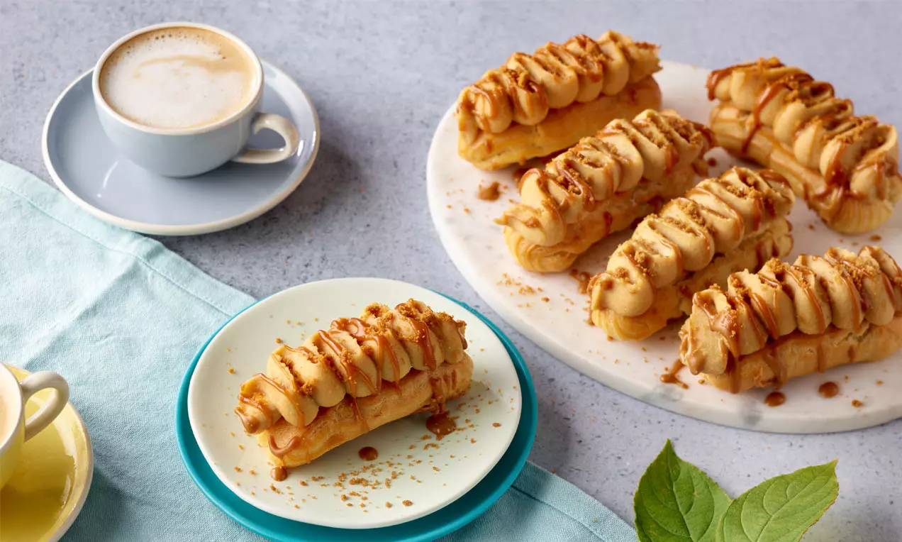 Lotus-Eclairs mit goldbraunem Brandteig, gefüllt mit beiger Creme, garniert mit Karamellsauce und Keksstreuseln auf Tellern angerichtet
