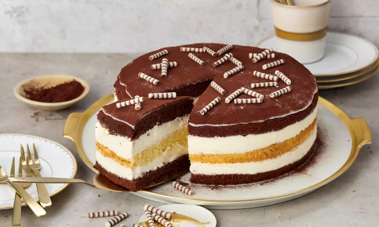 Torte mit dunkler Kakaoschicht, heller Cremefüllung und einem goldgelben Boden, dekoriert mit Schokorollen auf einer goldweißen Platte