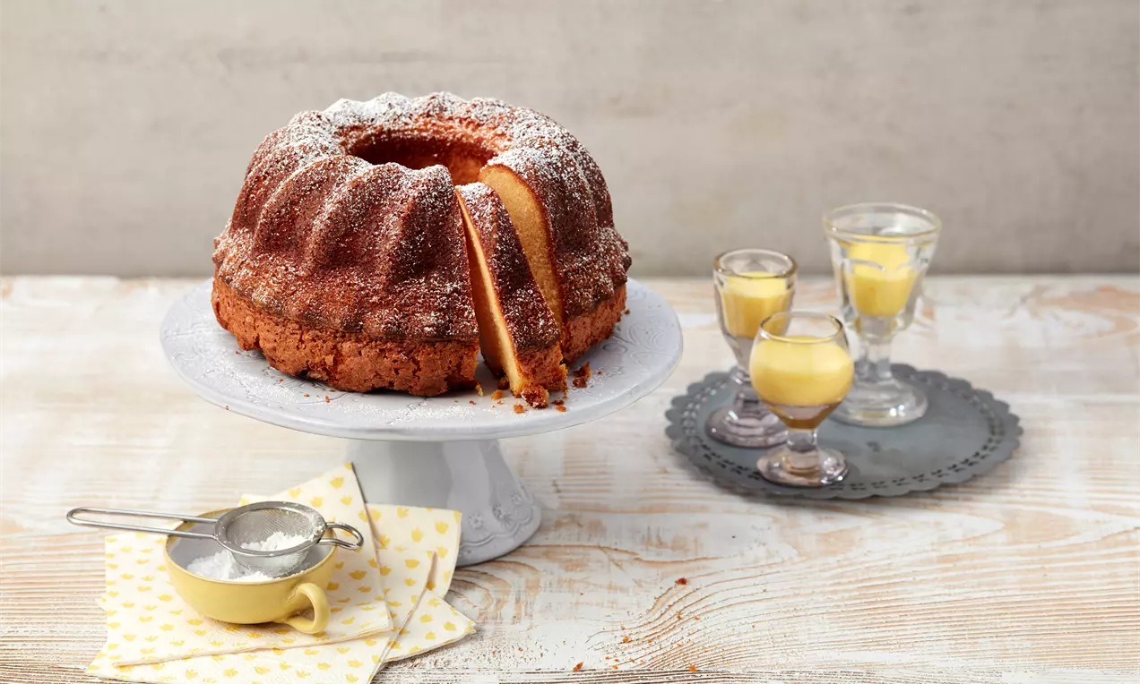 Goldbrauner Gugelhupf mit glatter Kruste, leicht bestäubt mit Puderzucker, serviert auf einer weißen Kuchenplatte mit Eierlikörgläsern