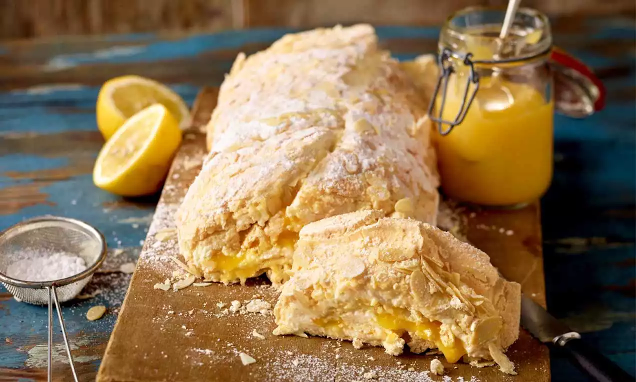 Gebackene Zitronen-Baiser-Roulade mit goldgelber Füllung auf Holzbrett, bestäubt mit Puderzucker, daneben Zitronen und ein Glas Zitronencreme