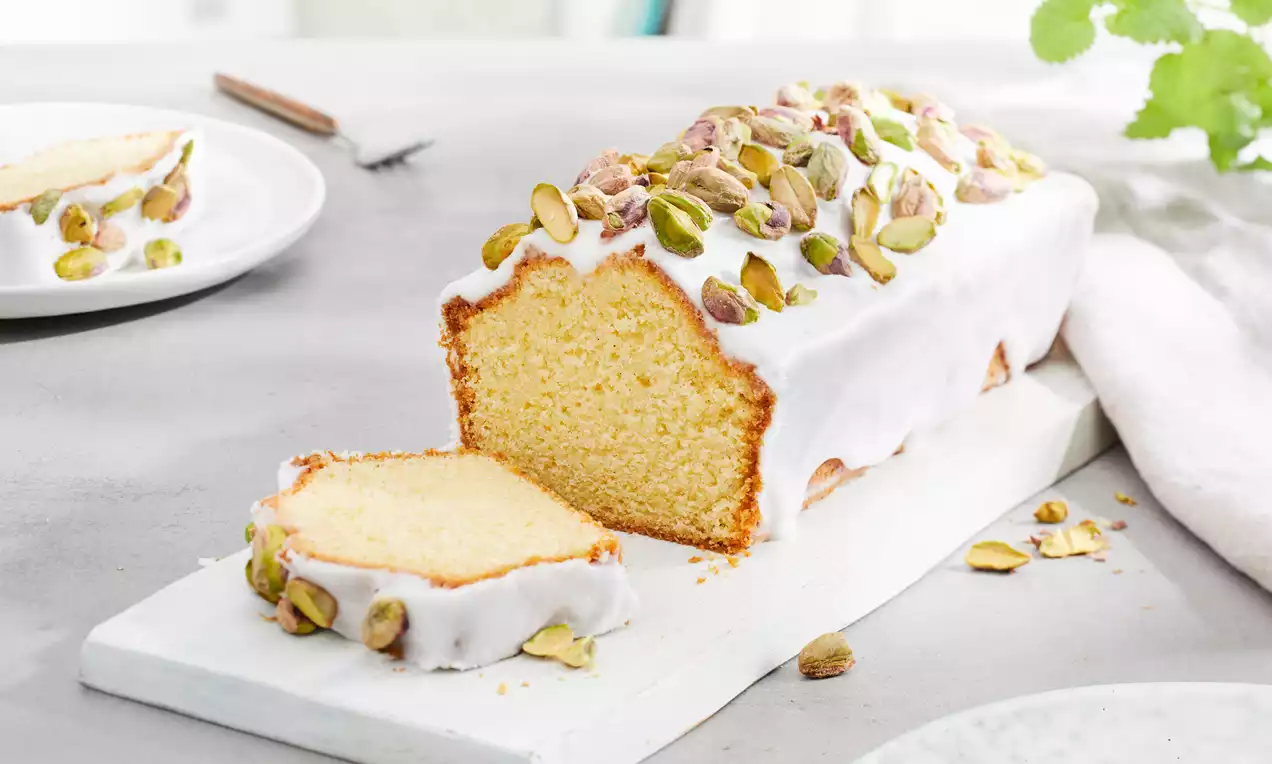 Gâteau doré en forme de cake, recouvert d’un glaçage blanc et parsemé de pistaches vertes sur une planche blanche