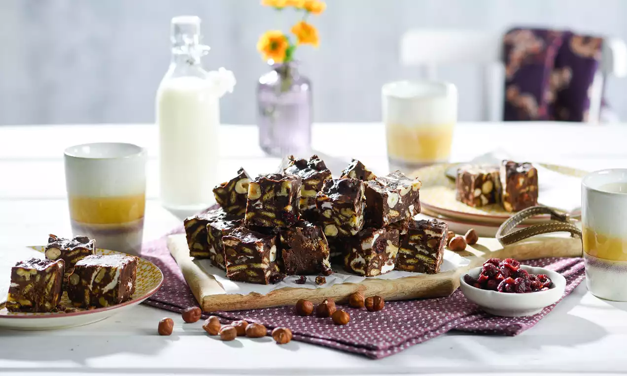 Rocky-Road-Stückchen mit dunkler Schokoladenbasis, Nüssen und Marshmallows auf Holzbrett, umgeben von Tassen, Milchflasche und Granatapfelkernen