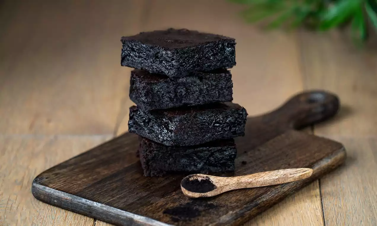 Tmavohnedé kocky brownies s vláčnou textúrou, naukladané na seba na drevenej doske vedľa malej drevenej lyžičky s kakaom