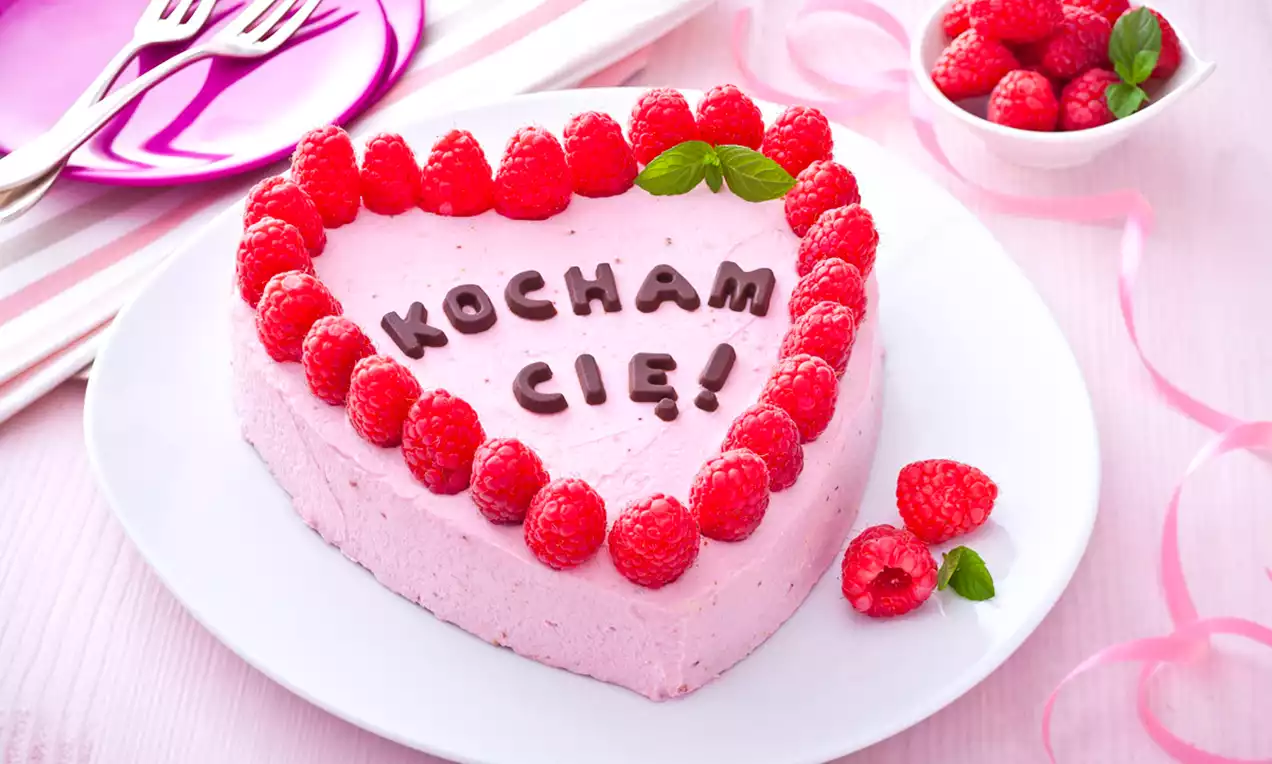 Serce z jasnoróżowego kremowego ciasta ozdobione malinami i czekoladowym napisem na białym talerzu, obok miseczka świeżych malin