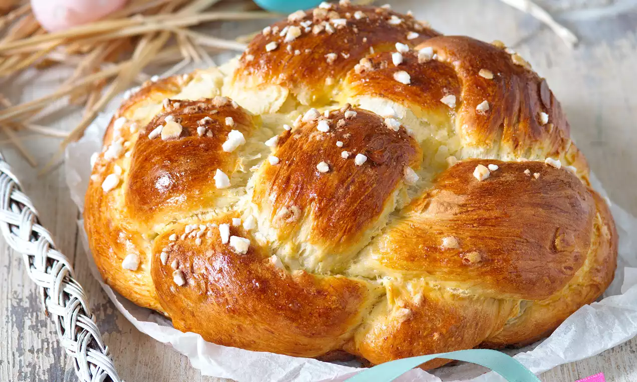 Lesklý dozlata upečený pletenec z mäkkého cesta, posypaný hrubým kryštálovým cukrom na papieri s jemnými stuhami a farebnými vajíčkami