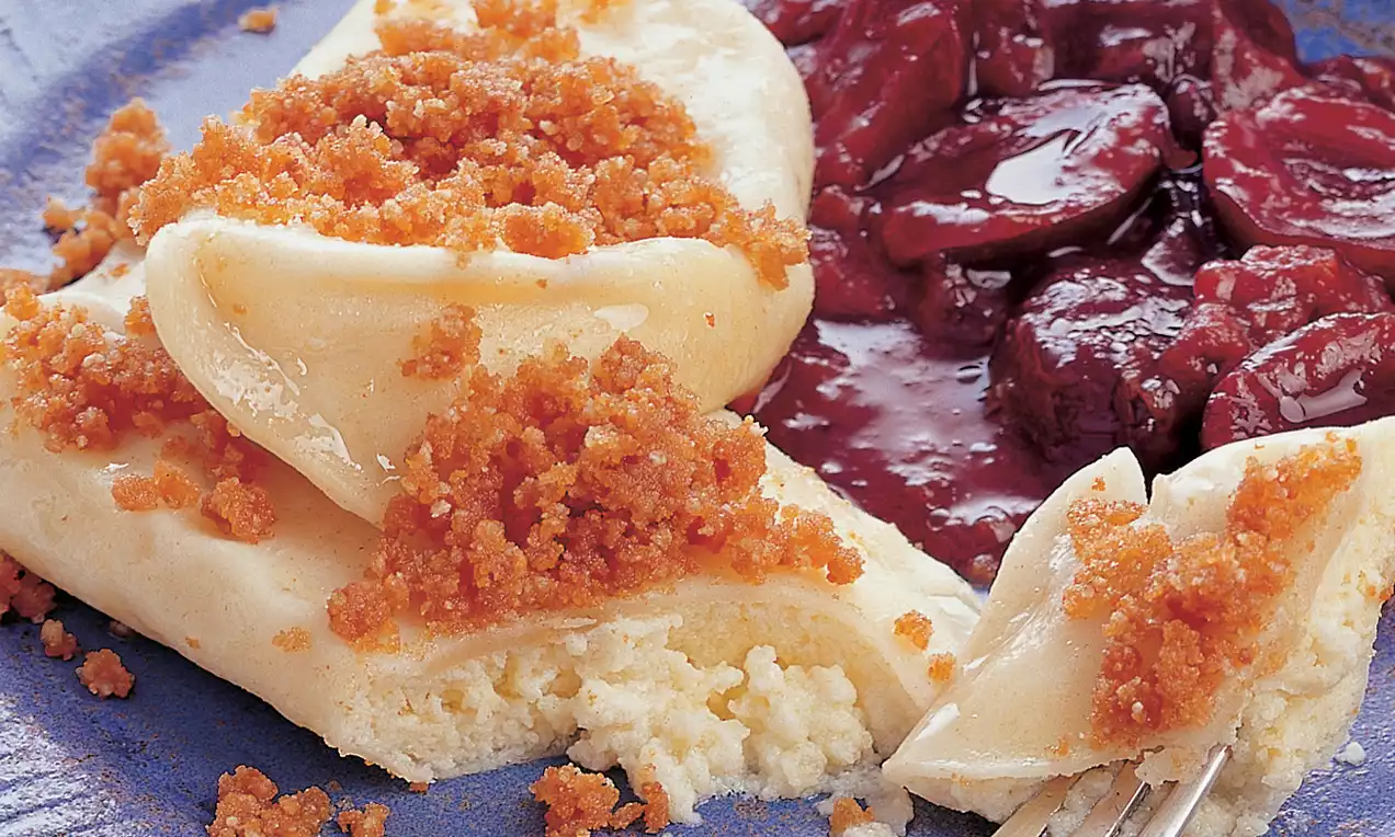 Strudelnudeln mit hellem Teig, goldbraunen Bröseln und cremiger Füllung, angerichtet neben glänzenden roten Kompottfrüchten auf einem blauen Teller