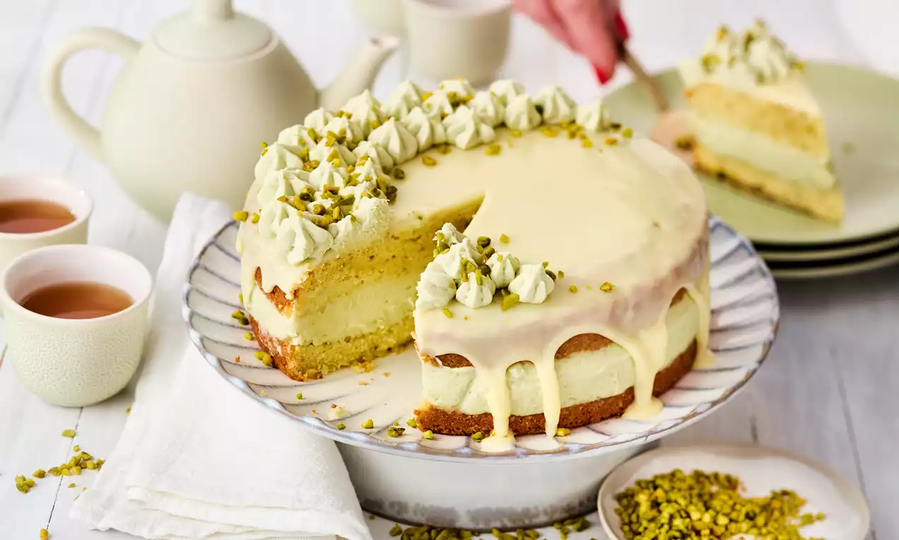 Pistazientorte mit hellgrüner Pistaziencreme, weißem Schoko-Drip, gehackten und halbierten Pistazien als Dekor auf weißem Teller