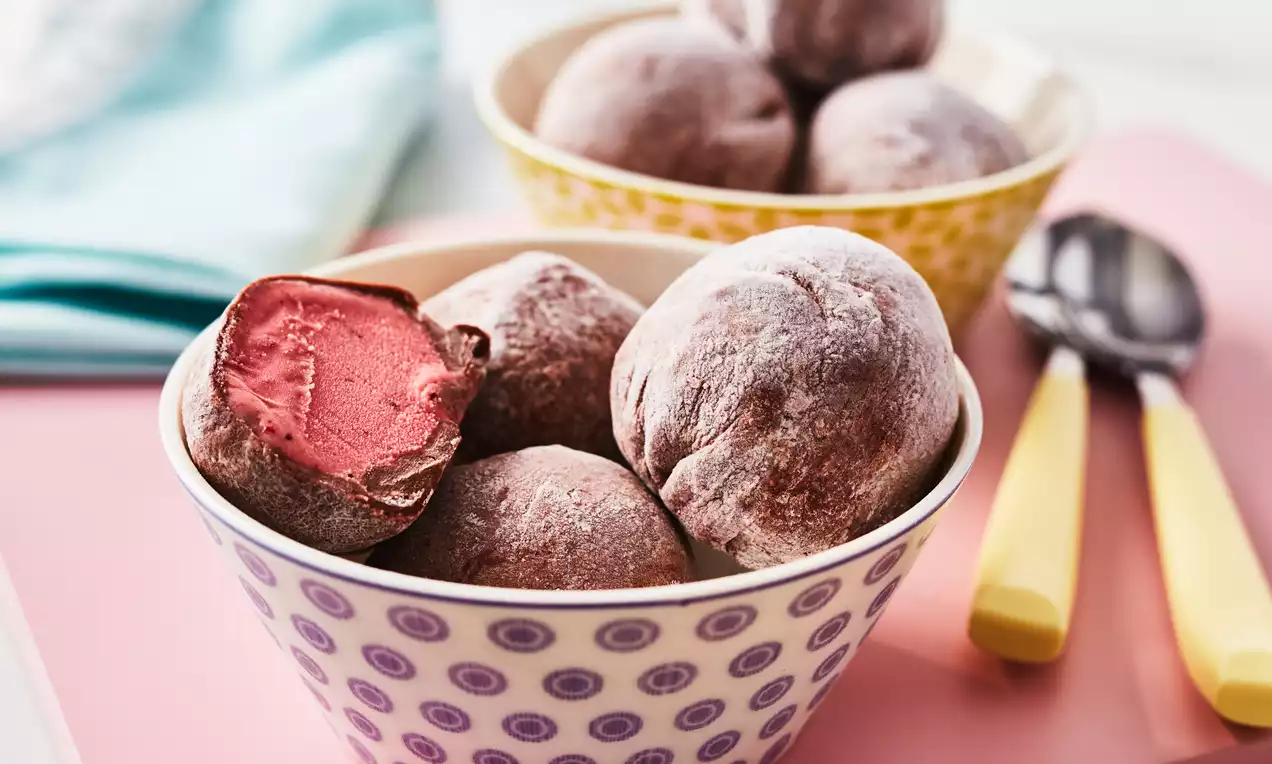 Soft round mochi balls dusted with pale white flour, deep red ice cream visible in a cut piece, arranged in patterned bowls on a pink surface