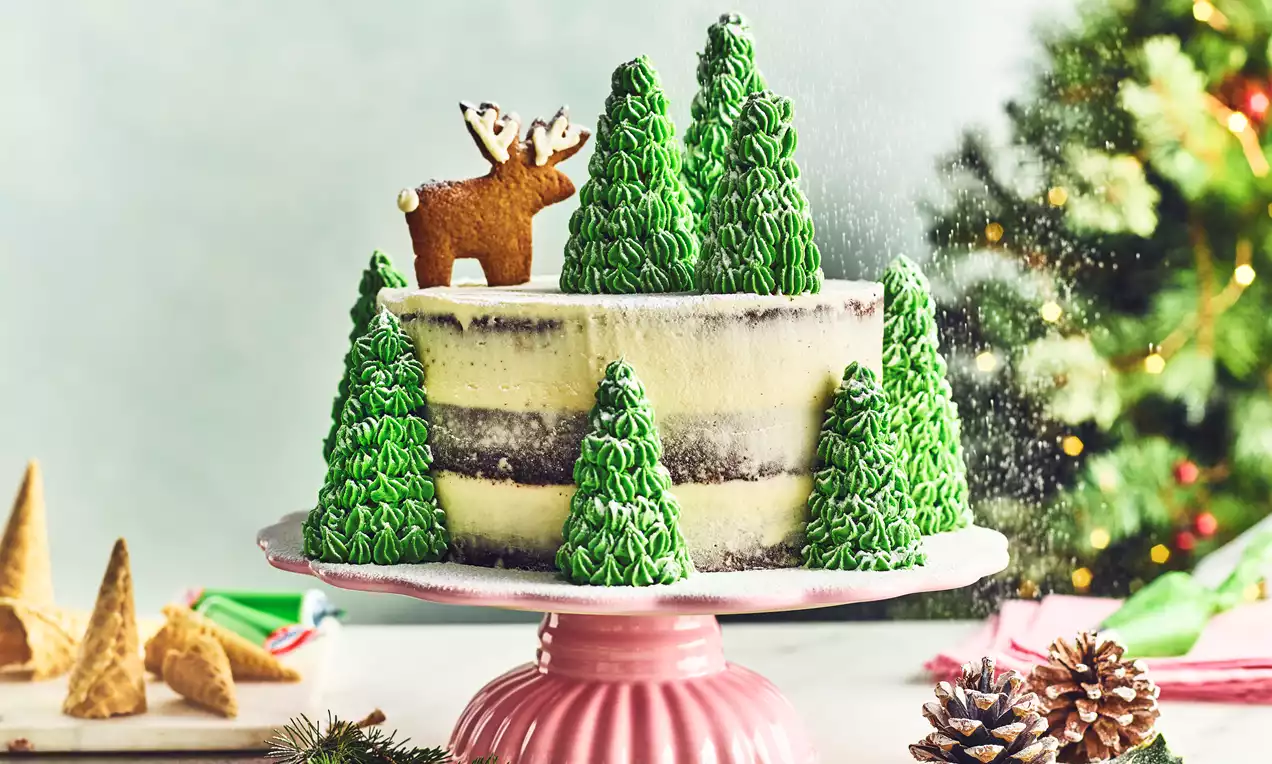 Gâteau à étages crème et chocolat orné de sapins en glaçage vert et d’un biscuit renne sur un présentoir rose avec des décorations de Noël