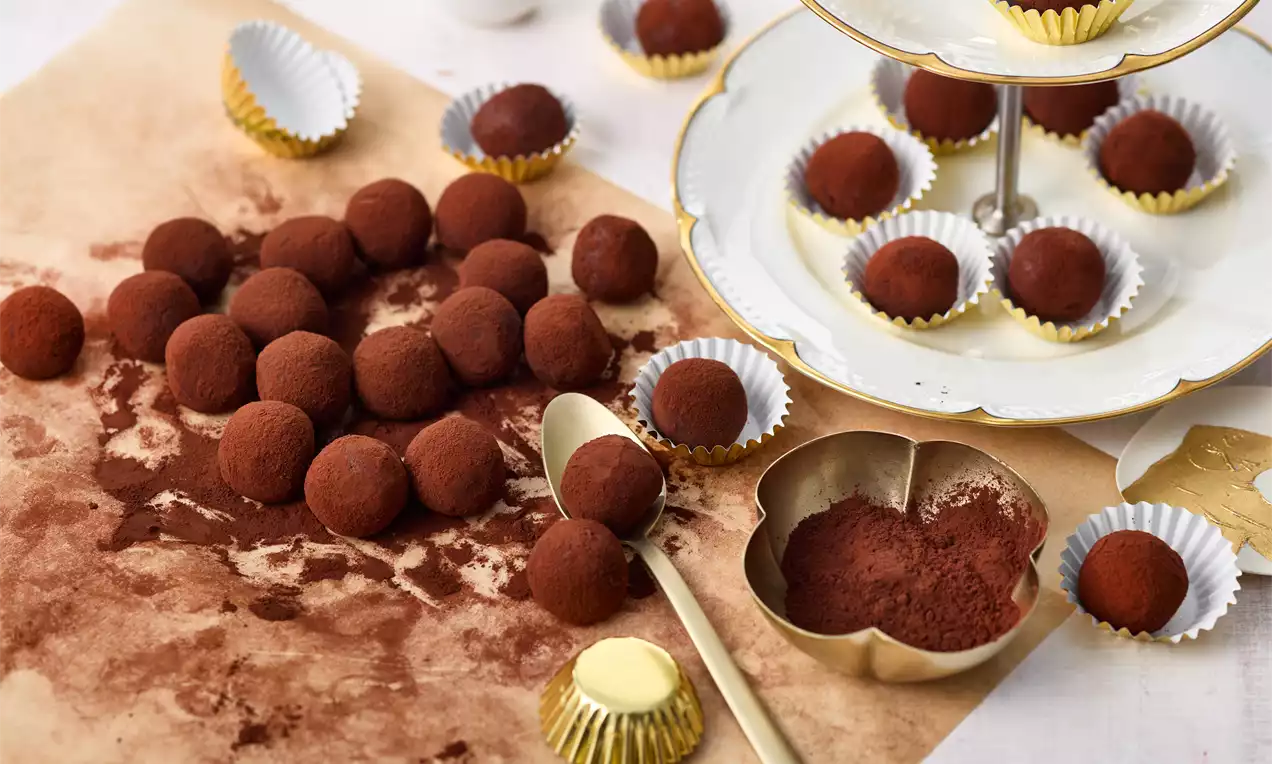 Kleine, kugelige Tiramisu-Pralinen mit feiner Kakaoschicht auf braunem Pergament, daneben goldene Förmchen und ein Servierständer