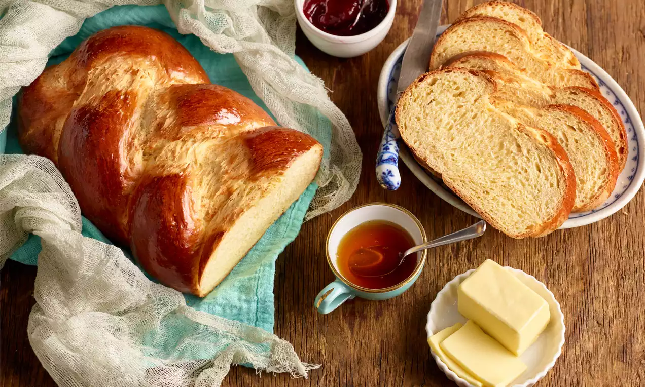 Glänzend brauner, locker geflochtener Germzopf mit weicher, hellgelber Krume neben Butterstücken, Honig und Marmelade auf Holzoptik