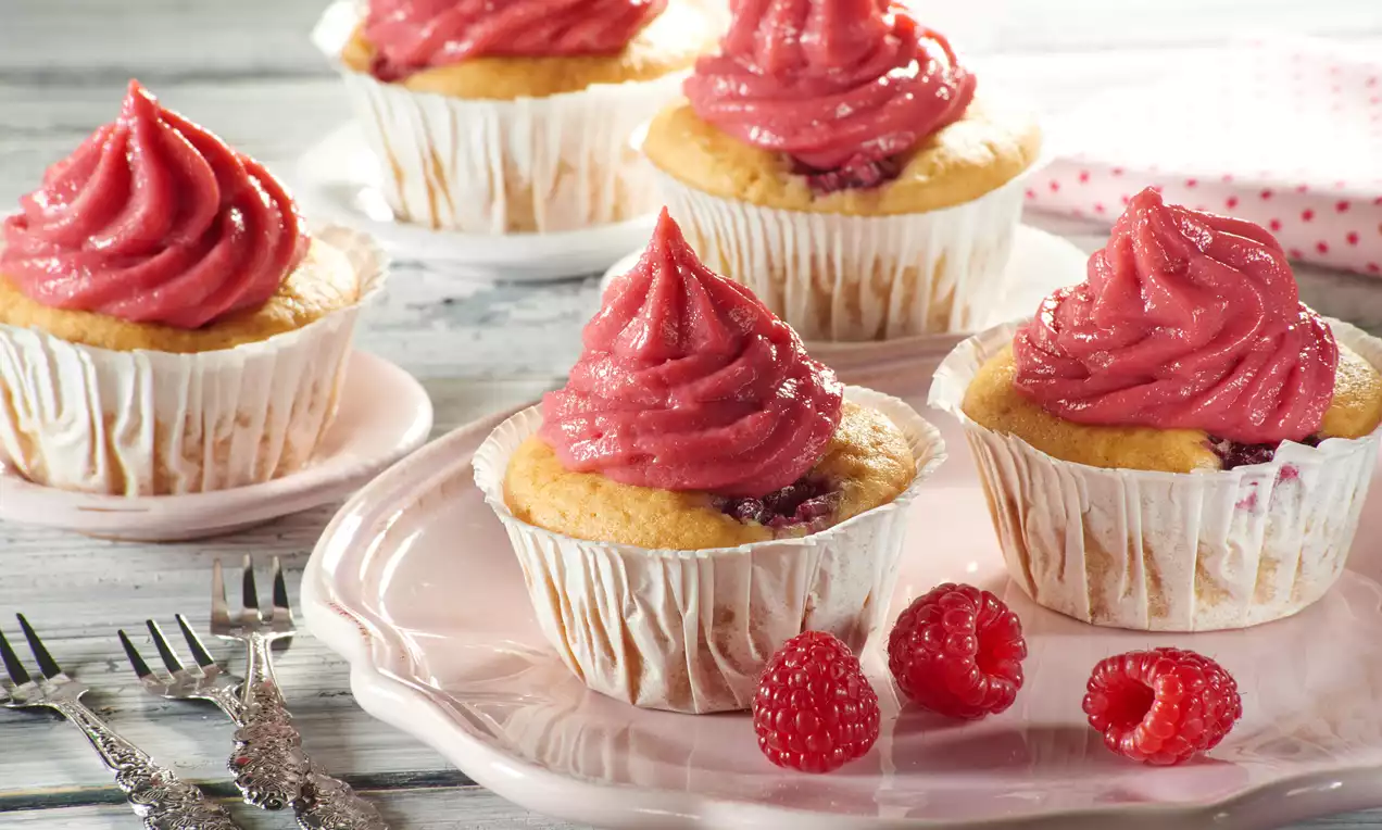 Vegane Himbeer-Cupcakes mit goldgelbem Teig, knallrosa Cremehaube in Papierförmchen auf rosafarbenem Teller mit frischen Himbeeren drapiert
