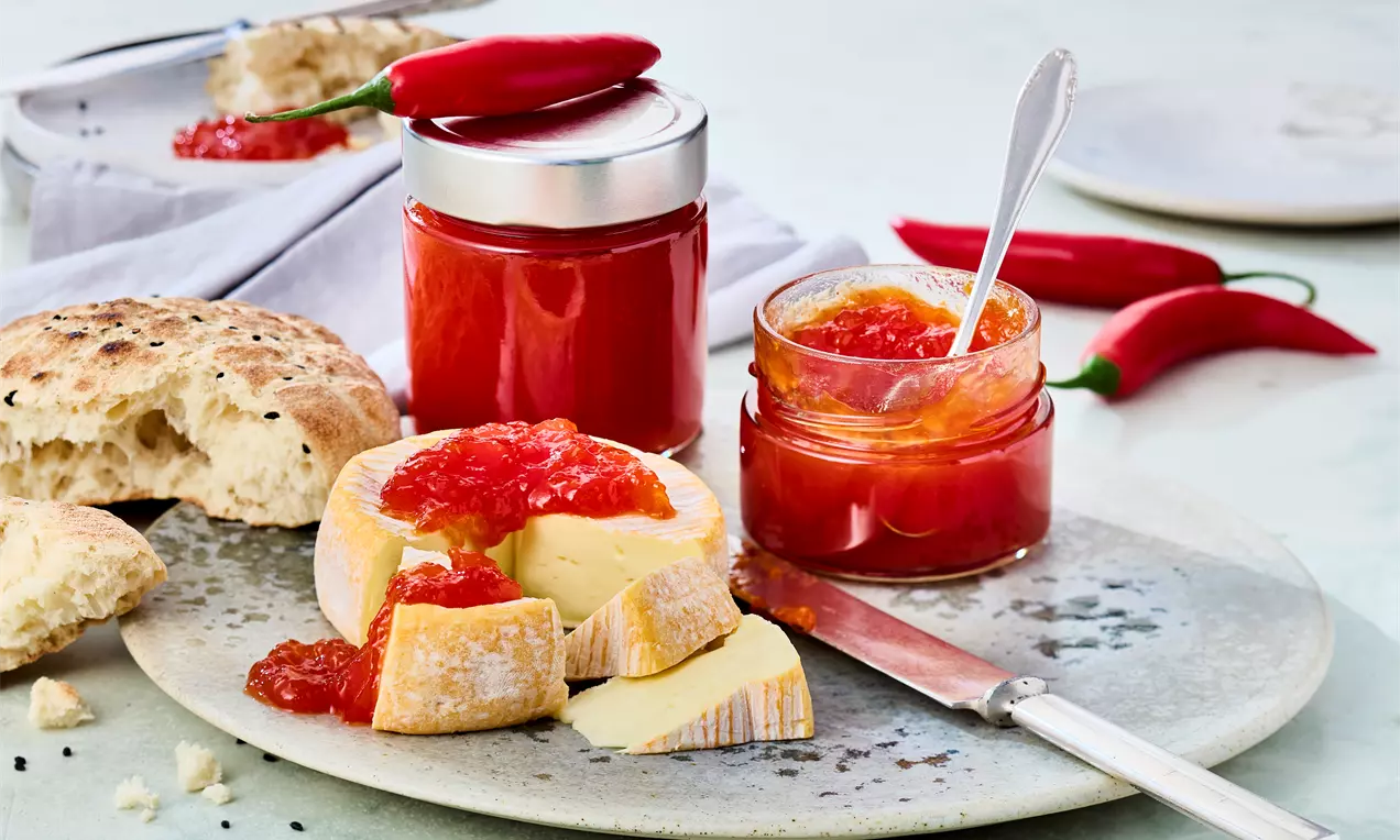 Chili Marmelade im Glas mit glänzender, tiefroter Masse und sichtbaren Chilistücken, Löffel daneben, serviert mit Brot und roten Chilis