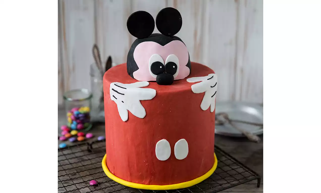 Torta cilindrica rossa con dettagli bianchi, decorata con testa tridimensionale di Mickey Mouse nero e rosa e guanti bianchi applicati