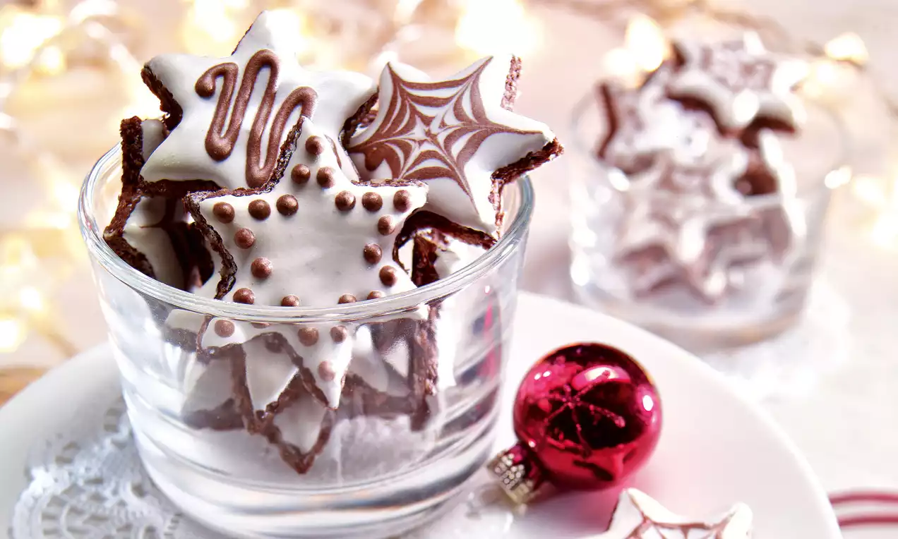 Zvjezdasti kolačići tamnosmeđe podloge s bijelom glazurom i čokoladnim uzorcima u staklenoj posudi na bijelom tanjiru s crvenom kuglicom