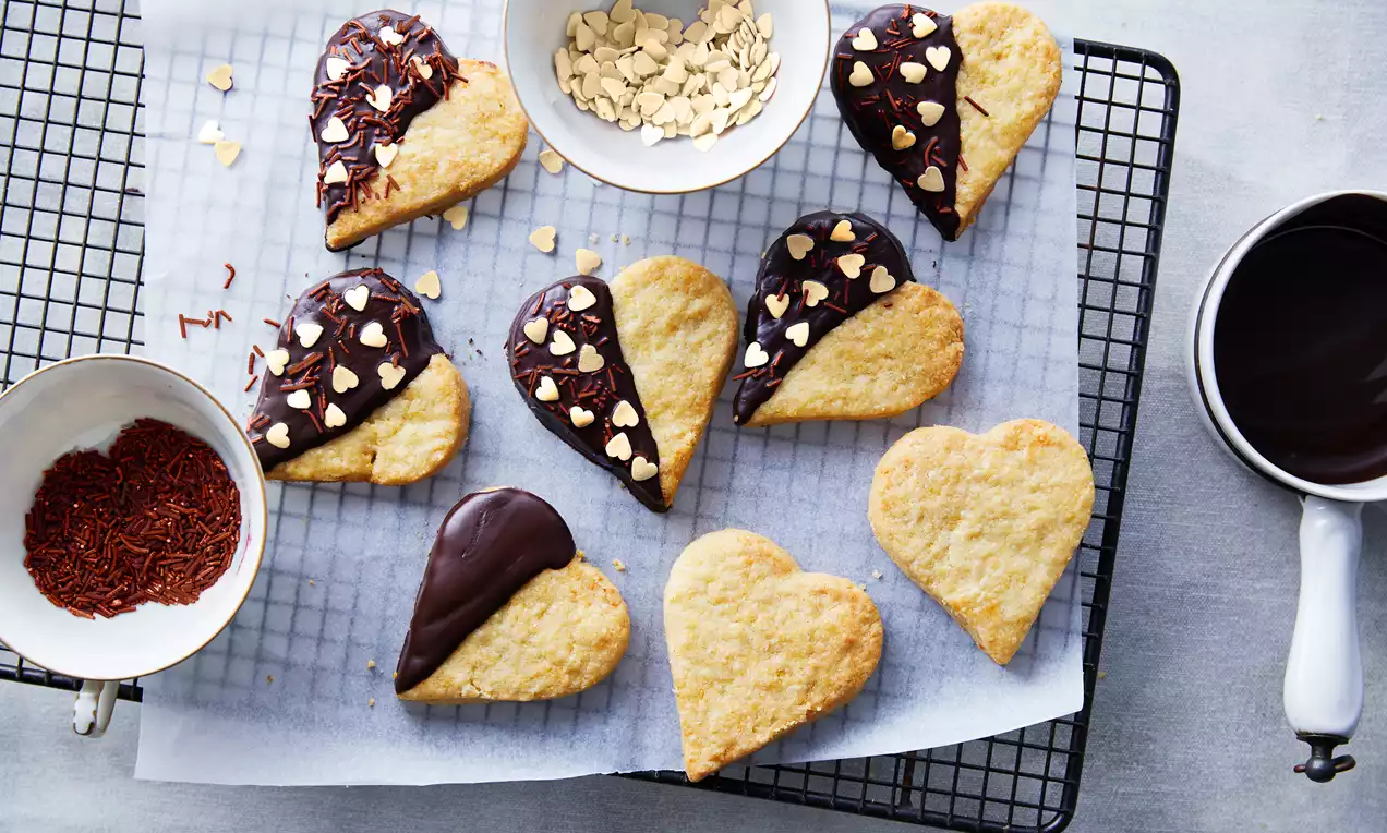 Harten koekjes met een lichtgele, kruimelige textuur, deels gedipt in donkere chocolade, versierd met hartjes en amandelschaafsel op een rek