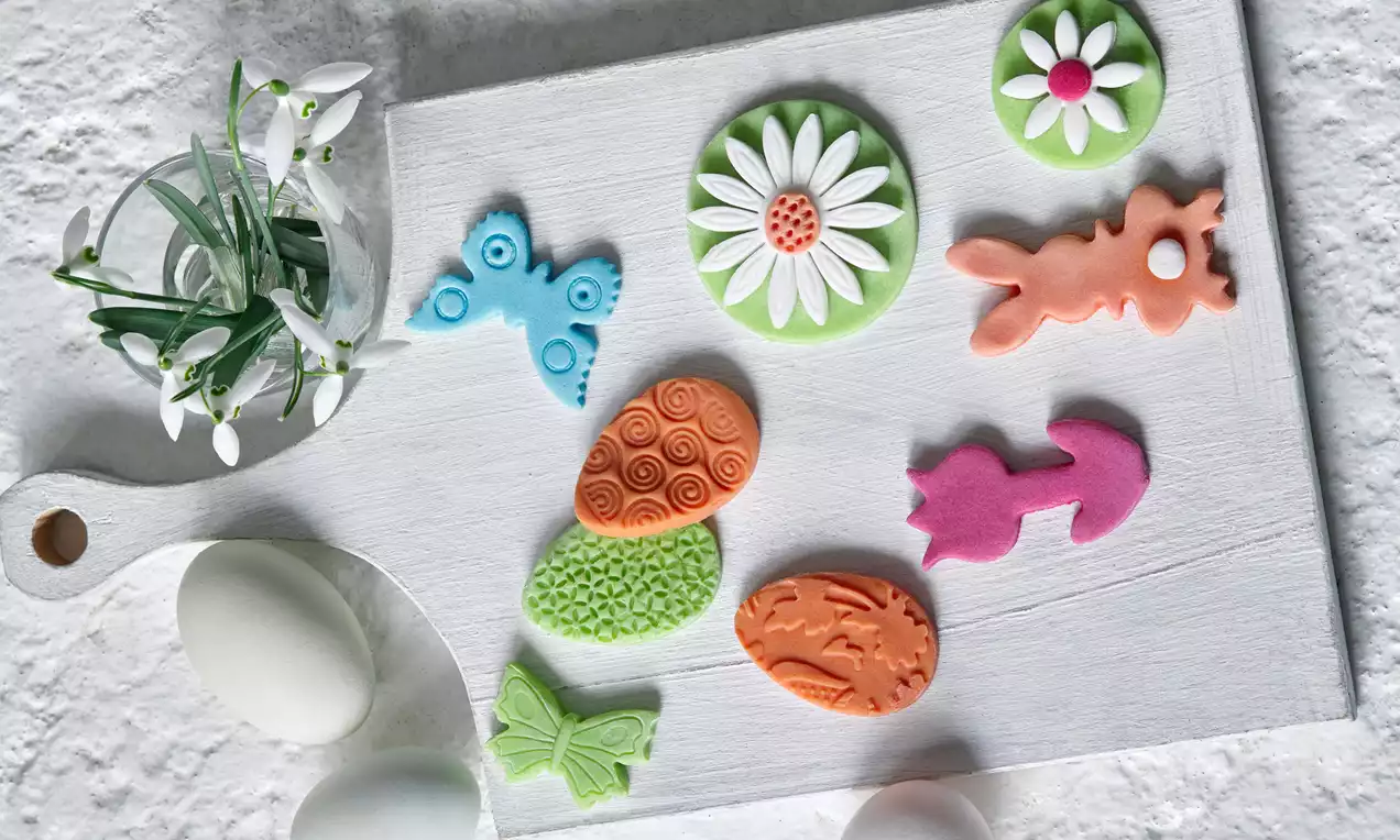 Farebné jarné dekorácie v tvare motýľov, kvetov, zajačikov a vajíčok na bielom drevenom podnose, vedľa bielych vajec a snežienok