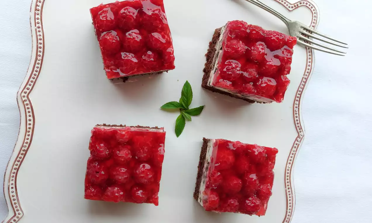 Čtvercové kousky dortu s vrstvou tmavého korpusu, světlého krému a lesklých malin, uspořádané na ladícím talíři s lístky máty