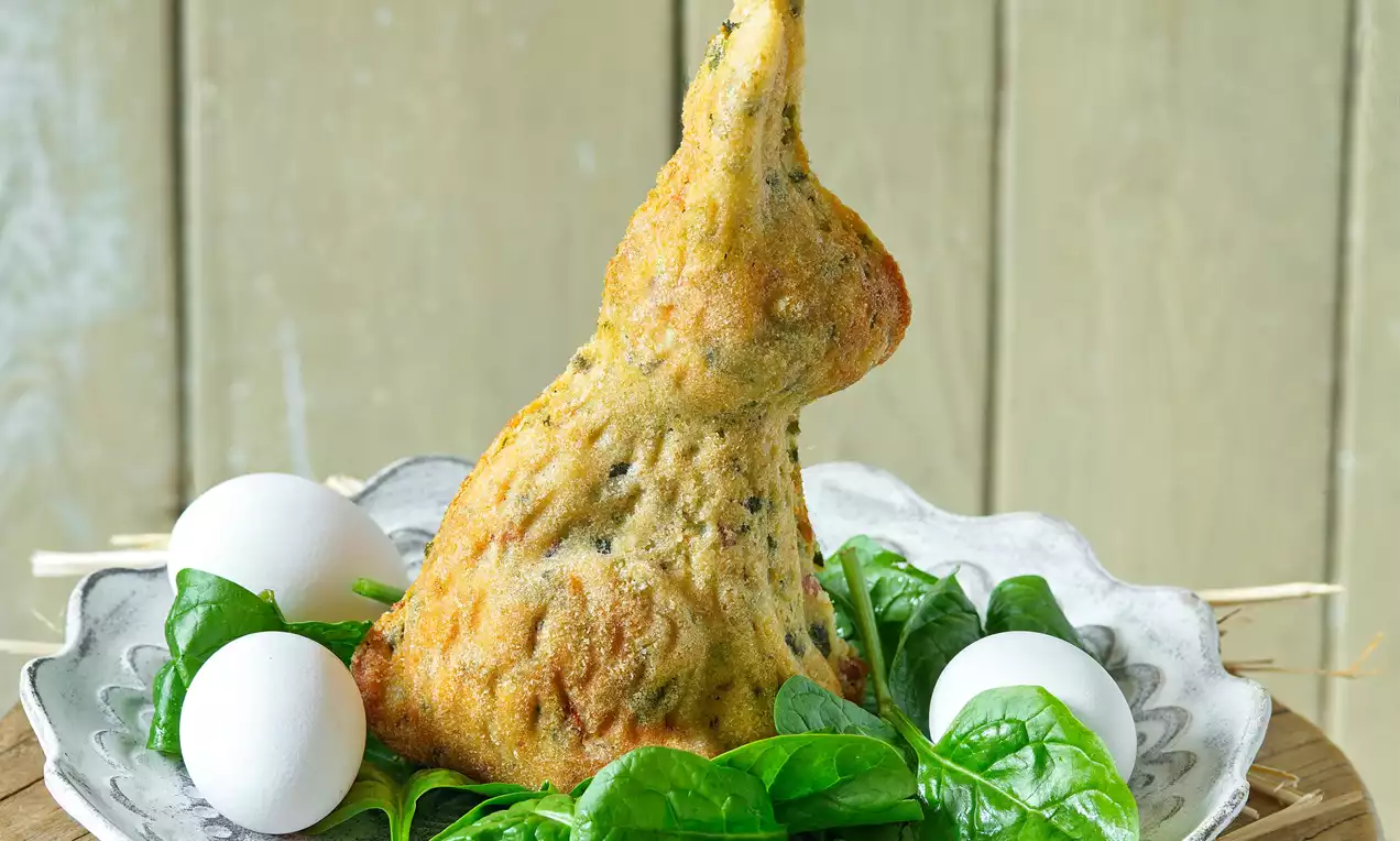 Zlatistý slaný zajačik v tvare sediaceho králika s jemnou textúrou, obklopený čerstvým špenátom a bielymi vajíčkami na ozdobnom tanieri