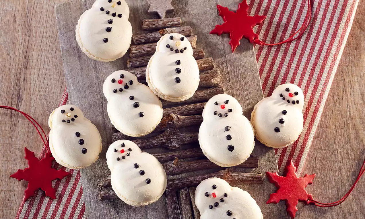 Krémově bílé makronky ve tvaru sněhuláků s černými knoflíky a úsměvy, uspořádané na dřevěné stromové dekoraci s červenými hvězdami