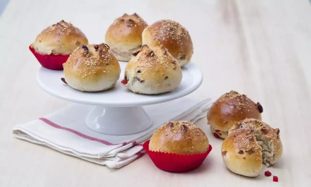 Zlatavě hnědé kulaté bochánky s křupavou kůrkou, posypané sezamem, na bílém stojanu a látce, některé v červených košíčcích, s rozinkami uvnitř