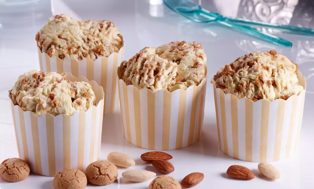 Vier cremefarbene Amaretto-Muffins mit bräunlicher Textur in gelb-weiß gestreiften Papierförmchen, dekoriert mit Mandeln und Amarettini
