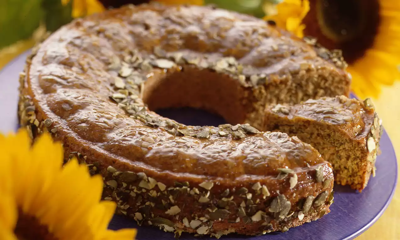 Goldbrauner Gugelhupf mit eingebackenen Kürbiskernen auf lilafarbenem Teller, umrahmt von leuchtend gelben Sonnenblumen