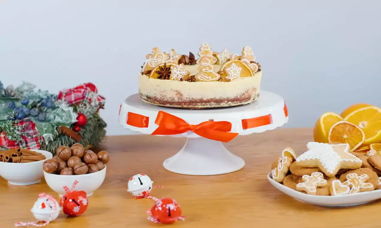 Vysoký krémový cheesecake na bielom stojane s červenou stuhou, ozdobený perníčkami v tvare hviezd a stromčekov s bielou polevou