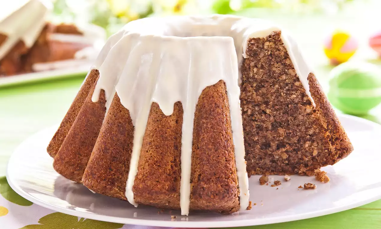 Babka orzechowa w formie wieńca z jasnobrązowym wnętrzem, oblana białą polewą na białym talerzu, otoczona kolorowymi dekoracjami