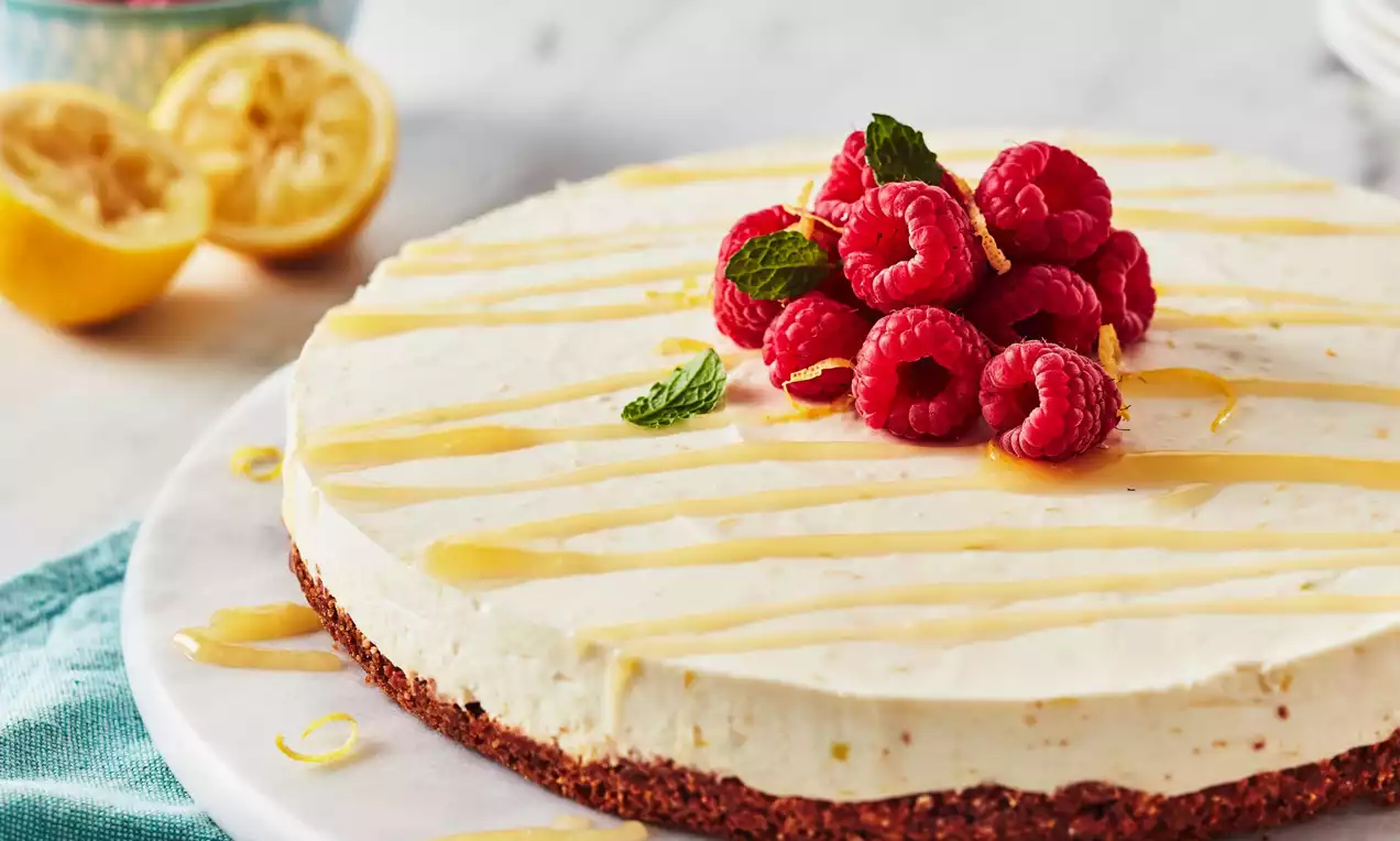 Pale yellow cheesecake with smooth creamy texture on a brown biscuit base, topped with raspberries, mint leaves and lemon drizzle on a white plate
