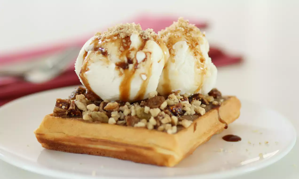 Altın rengi waffle üzerinde beyaz dondurma topları, tahin ve pekmez sosuyla süslenmiş, kıyılmış cevizlerle tamamlanmış beyaz tabakta