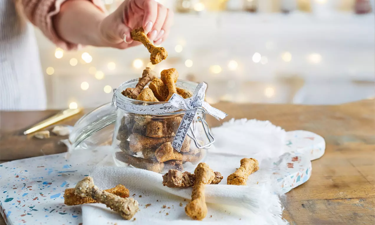 Golden-brown bone-shaped dog biscuits with a crumbly texture spilling from a glass jar tied with ribbon onto a white cloth