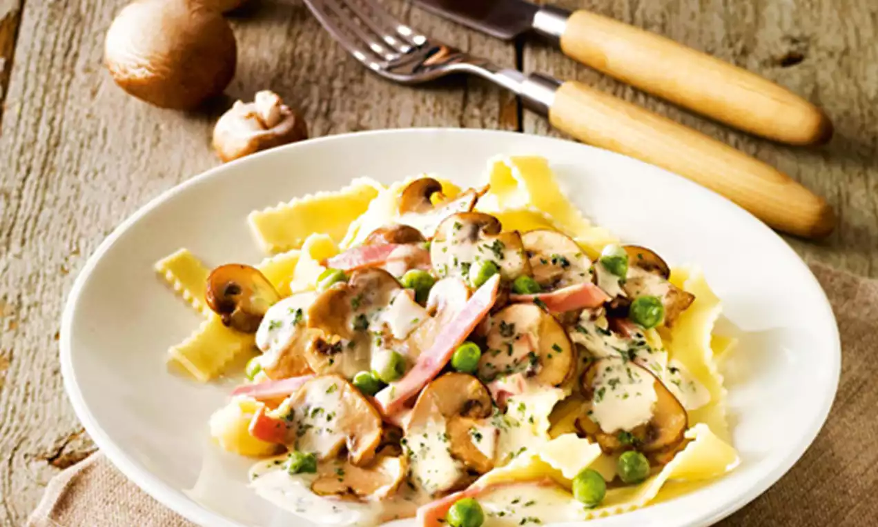 Gelbe Bandnudeln mit cremiger Pilz-Sahnesoße, Schinkenstreifen und grünen Erbsen auf weißem Teller mit rustikalem Holzhintergrund