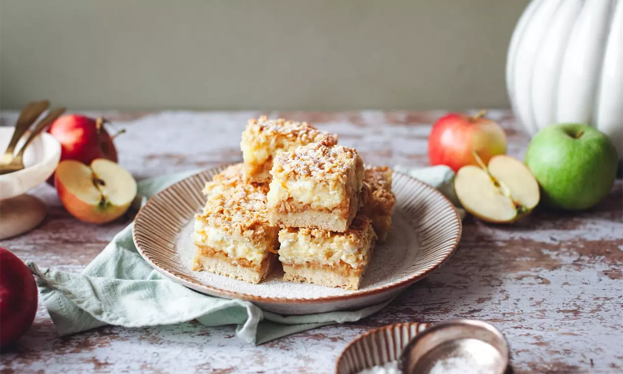 Štvorcové kúsky jablkového koláča so zlatistou strúhankovou posýpkou na vrchu, uložené na béžovom tanieri obklopenom jablkami a dekoráciami
