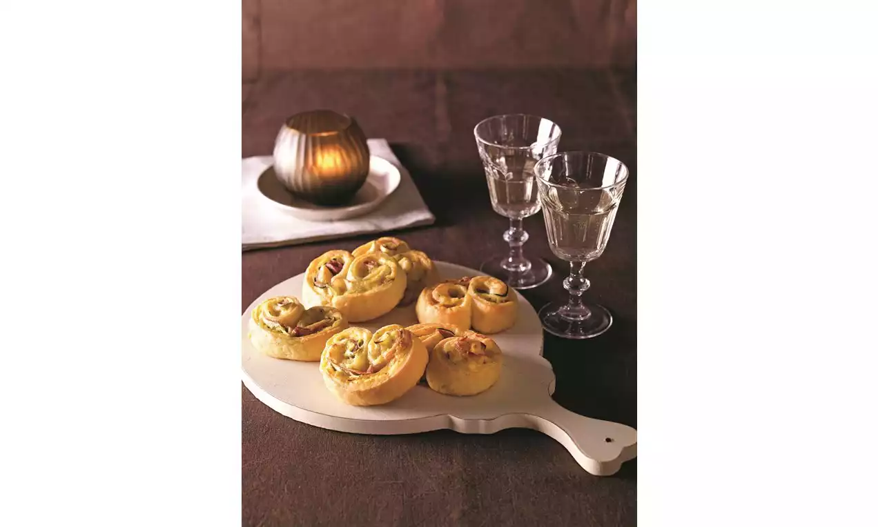 Zlatisté špirálky z nadýchaného cesta s kúskami šunky a cukety na drevenom lopáriku, vedľa sklených pohárov a sviečky na hnedom obruse