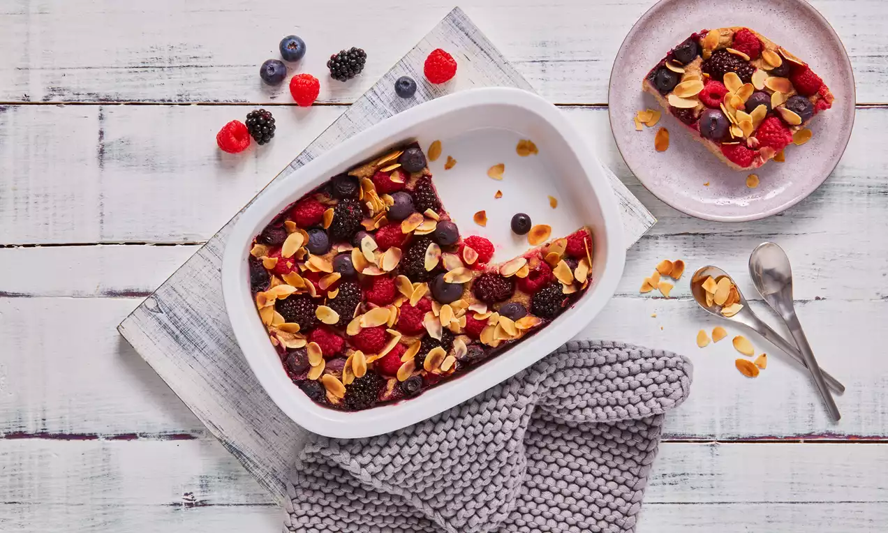 Goldbraune Baked Oats mit frischen roten und dunklen Beeren sowie gerösteten Mandelblättchen in weißer Auflaufform auf hellen Holzbrettern