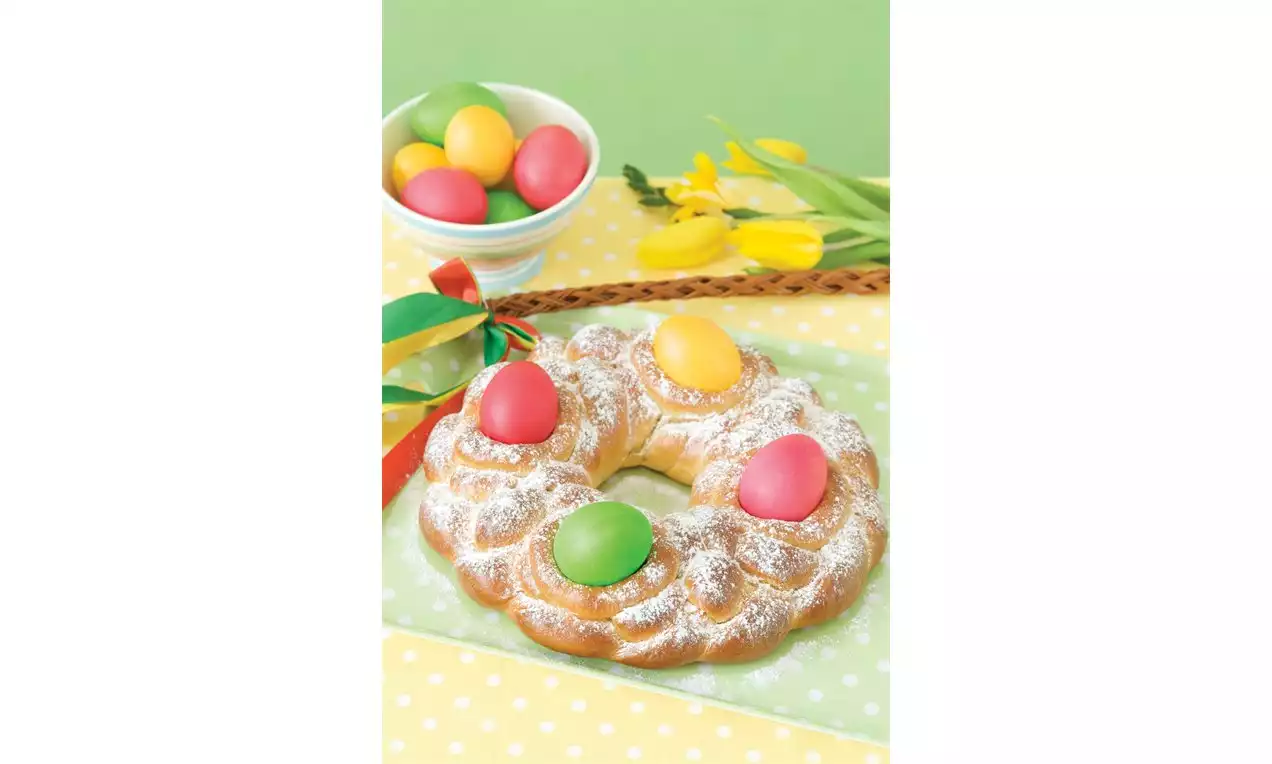 Velikonoční věnec ze zlatavého kynutého těsta posypaného moučkovým cukrem, zdobený barevnými vajíčky na puntíkatém žlutém ubrusu