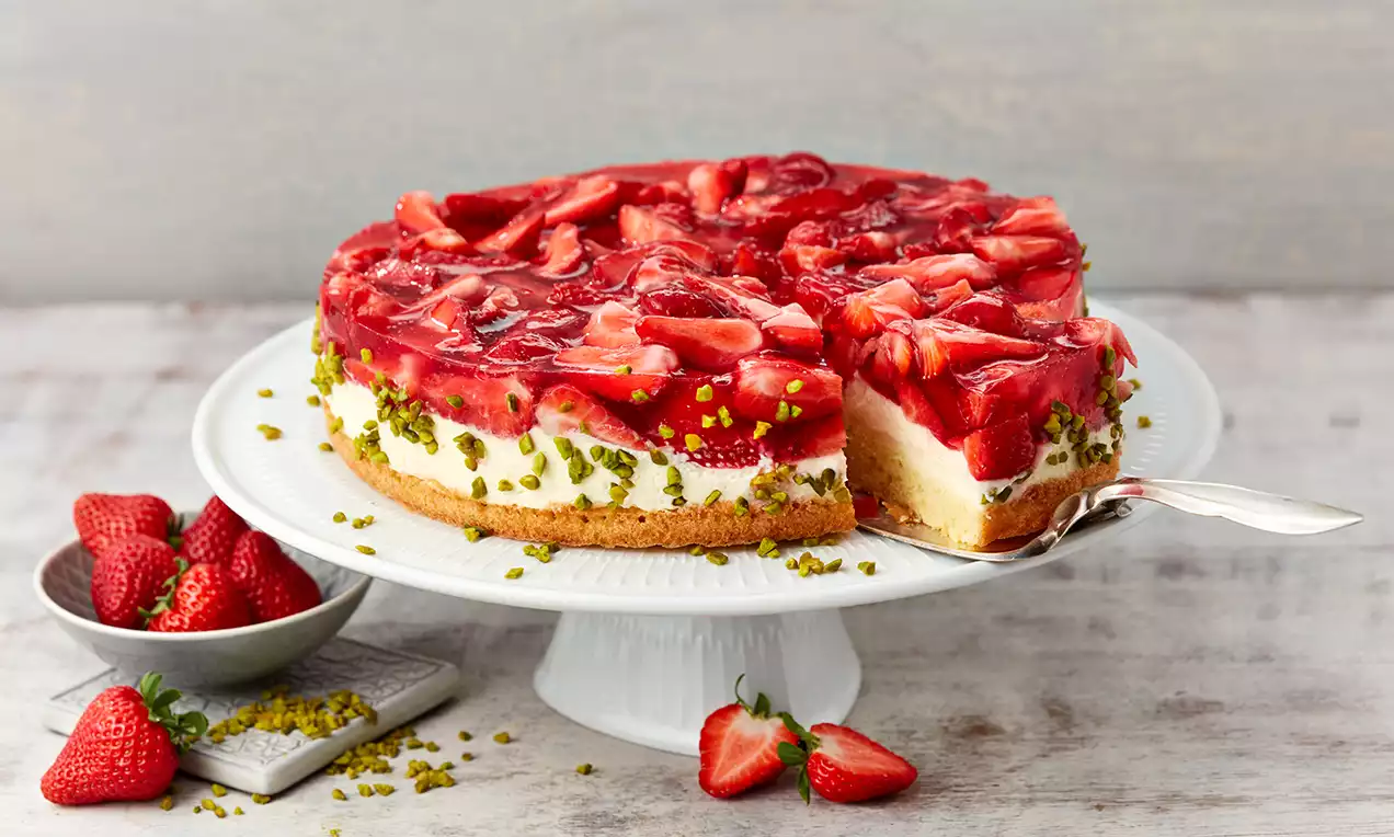 Torta od jagoda s laganom žutom biskvitnom podlogom, kremastim bijelim slojem i sjajnim crvenim preljevom od jagoda, ukrašena nasjeckanim pistaćima
