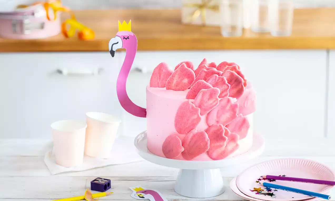Ronde roze cake op een wit taartplateau met suikerbloemblaadjes, flamingohalsdecoratie en een papieren kroon in een lichte keuken