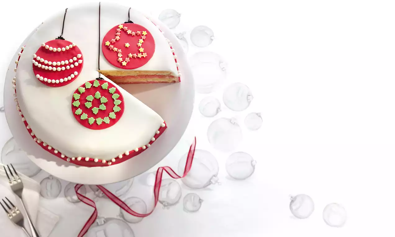 Runde Torte mit weißem Fondant, verziert mit roten Christbaumkugel-Motiven, ein angeschnittenes Stück zeigt eine helle Schichtfüllung