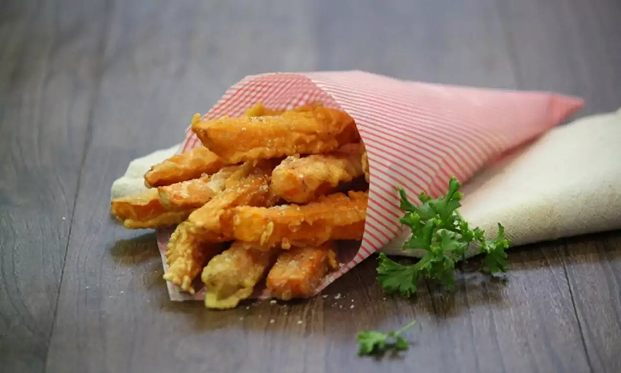 Golden orange sweet potato fries with a crisp texture spilling out of a red-striped paper cone, garnished with fresh parsley sprigs