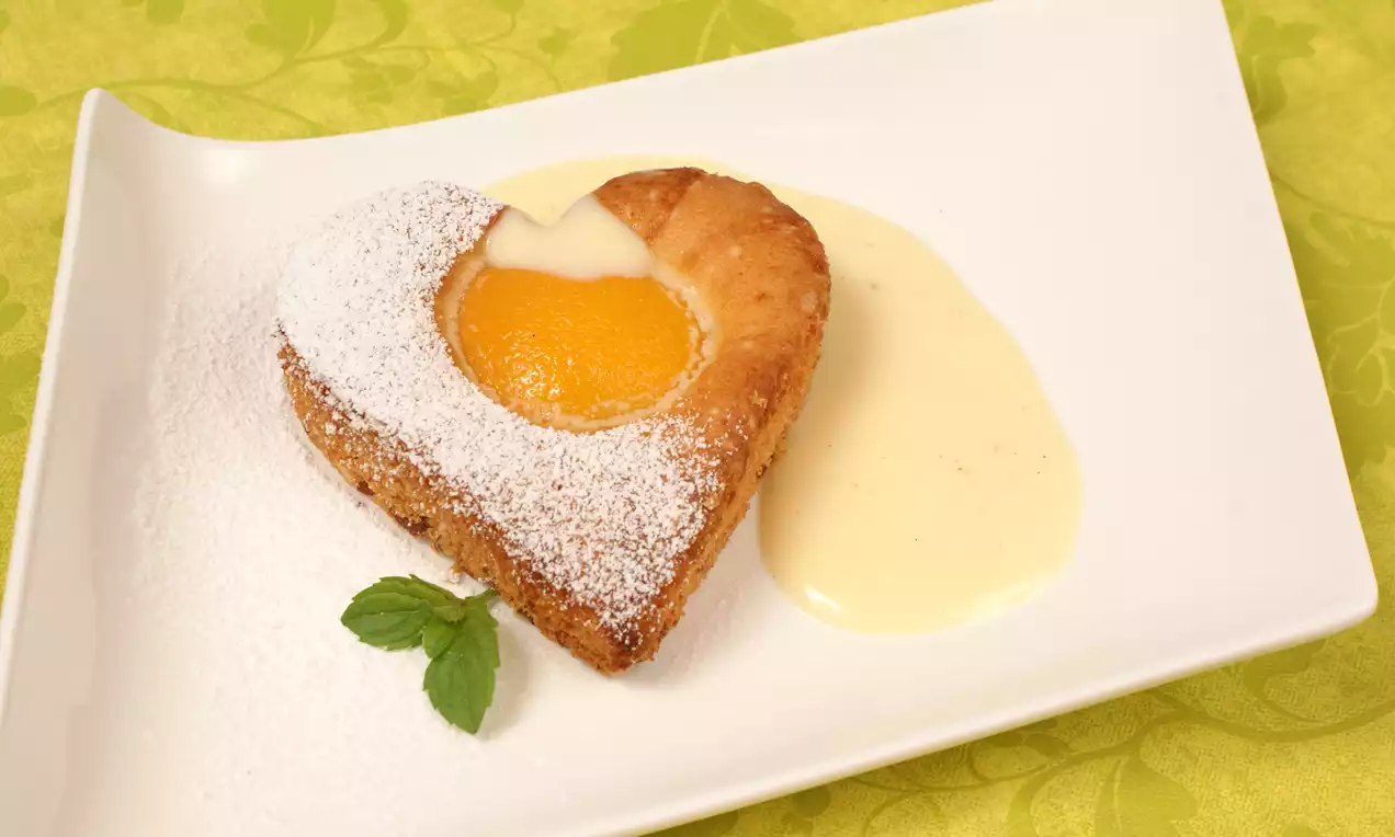 Goldbraunes, herzförmiges Gebäck mit saftigem Pfirsich und Puderzucker, serviert auf weißem Teller mit Vanillesauce und Minzblatt
